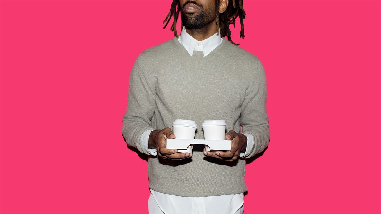 Stylish man with dreads and a beard holding two cups of to-go coffee against a hot pink background. 