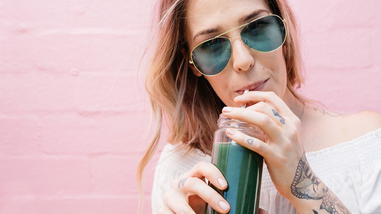 Young woman drinking a green juice with green sunglasses on pink background