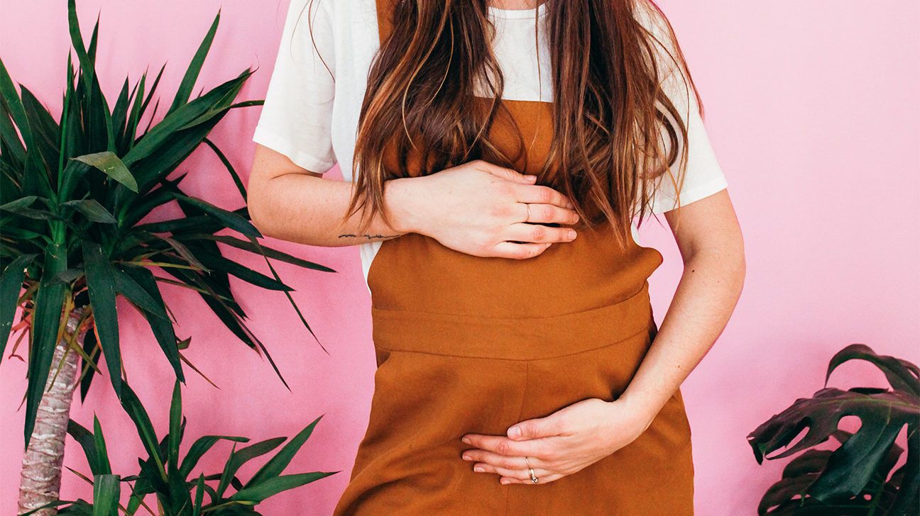 Pregnant woman wearing overalls in front of a pink background with palm plants.