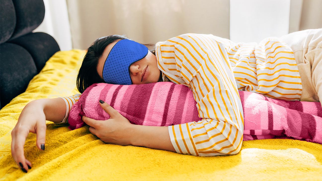 Image of heavy sleeping young woman lying on the bed with sleeping mask, having sweet dreams. Female lying on colorful pillow with blue sleep mask on her eyes resting at home header crop