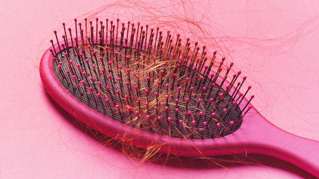 A bright pink hairbrush lying on a light pink background. There is red hair caught in the teeth of the hairbrush. 