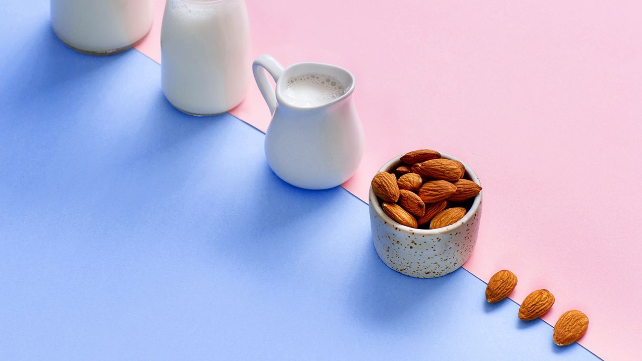 a clear jar and white pitcher of almond milk next to a dish of almonds on a pink and blue table top