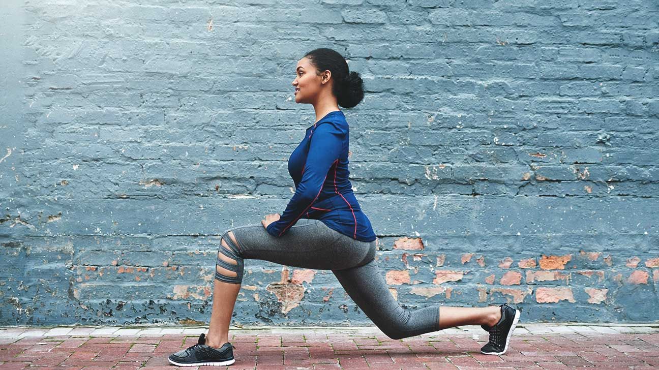 A woman doing a lunge stretch in front of a brick wall.