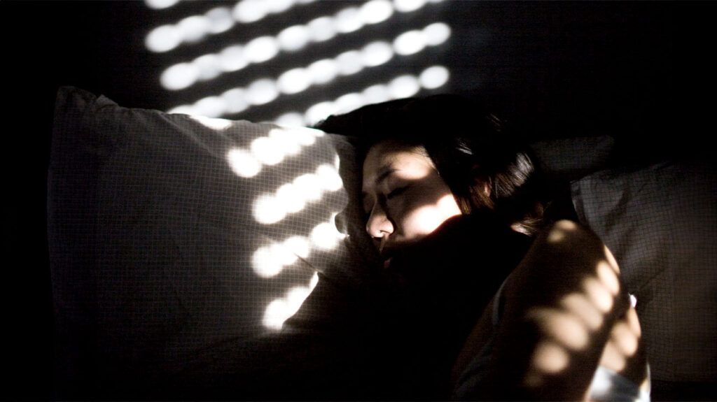 a woman asleep in bed with light from a window forming a pattern of dots on the wall behind her