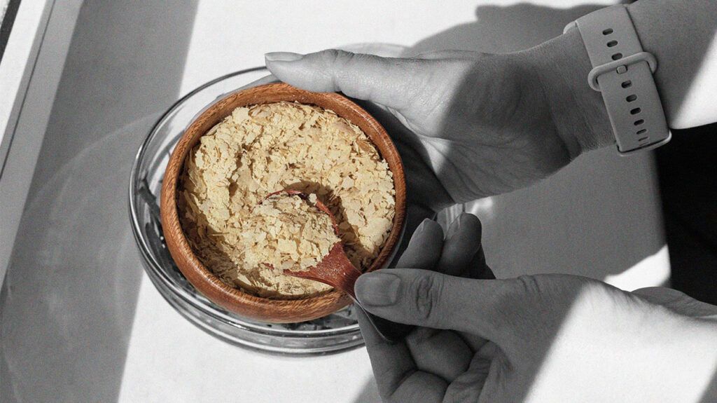 woman's hands scooping nutritional yeast flakes with a wooden spoon from a wooden bowl,