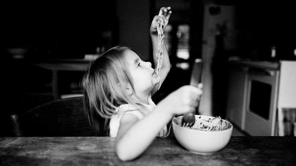 A little girl dangling spaghetti into her mouth at the dinner table.