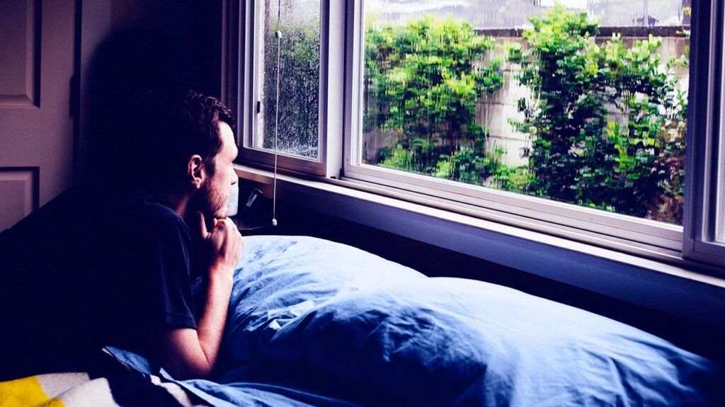 Man lying on bed staring through window at rain