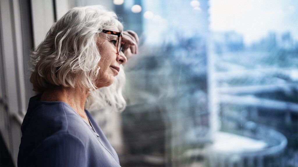 A worried older woman leaning against a window.