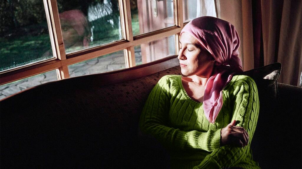 A woman with cancer is in a dark room sitting on a couch with her eyes closed