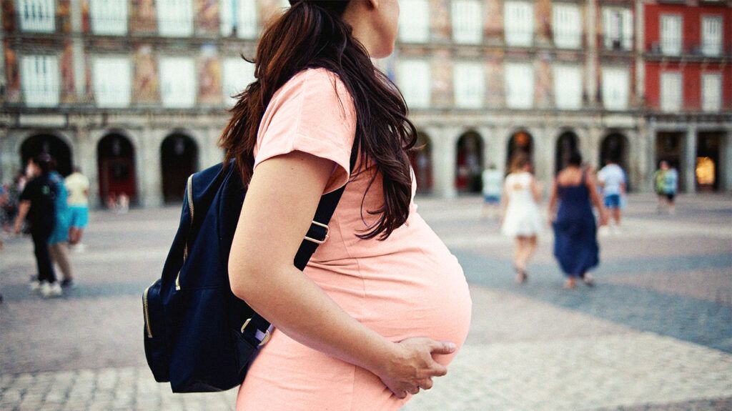 A pregnant person wearing a backpack, holding their stomach.-2