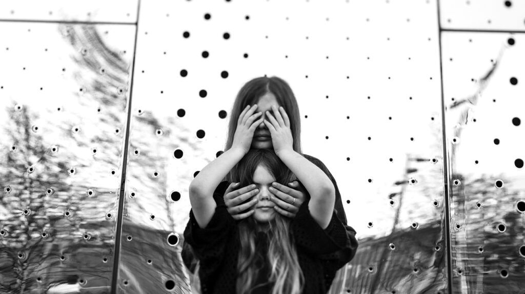Two girls covering their eyes in front of a shiny steel wall.