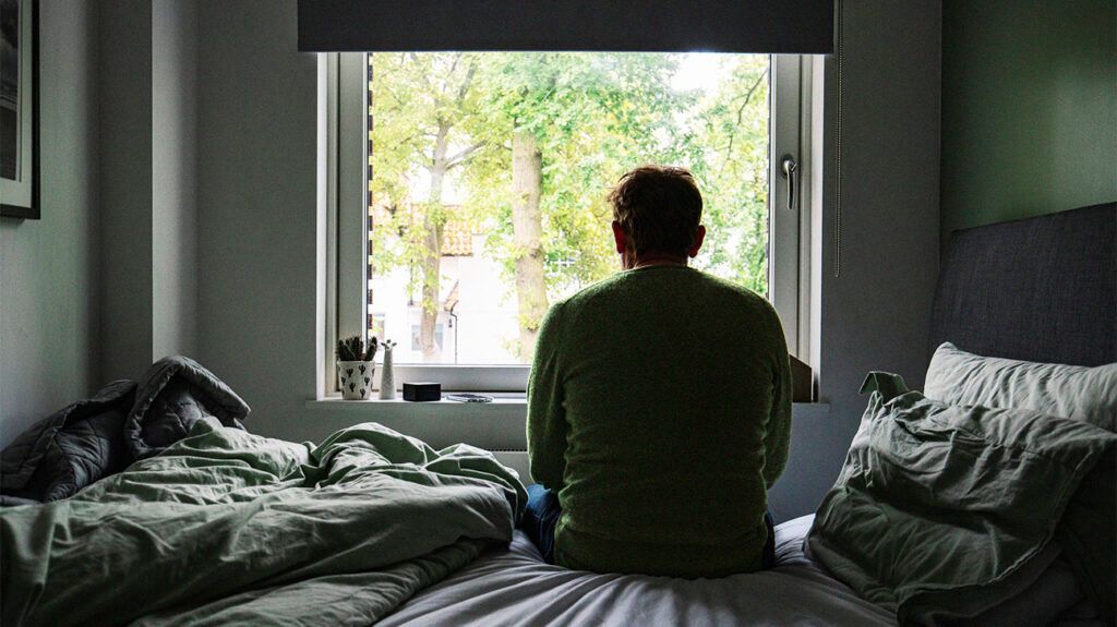 A person is sitting on a bed and looking out of a window.