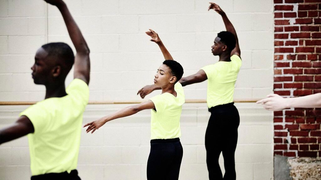 Three male ballerinas dancing. They are wearing yellow t-shirts.