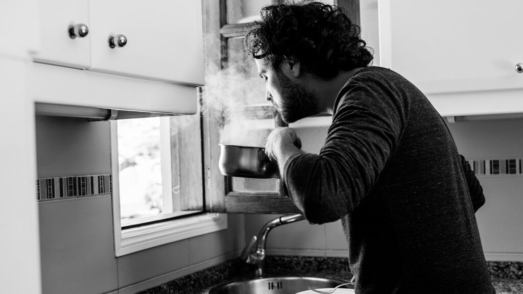 A young man at his kitchen sink smells the steam escaping from a pan.