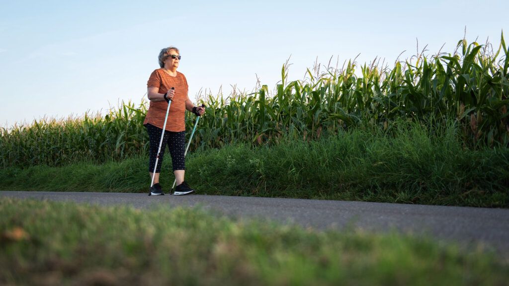 A woman using walking poles as she strides past a field of corn