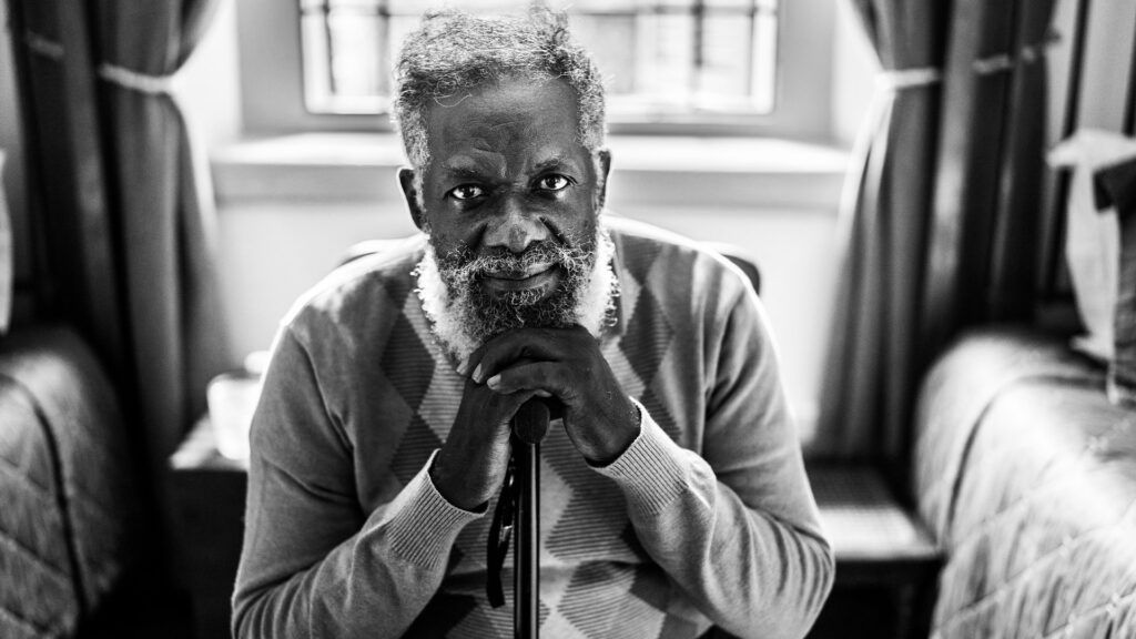 An older looks into the camera as he rests his chin on his cane while sitting