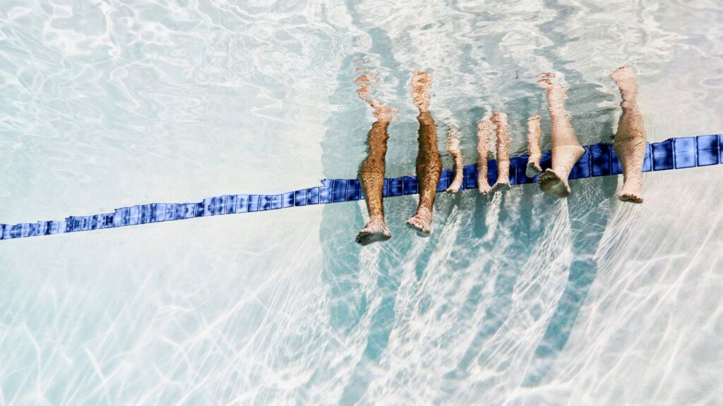 The feet of several people hanging into a swimming pool, them sitting above the water surface, out of view