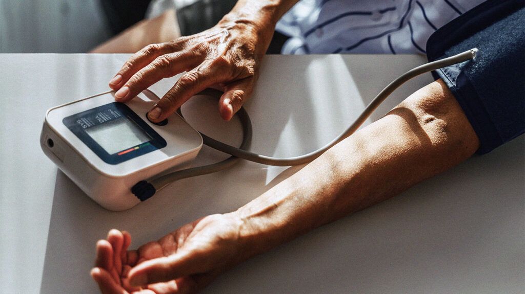 An older adult measures their blood pressure using a monitor with their arm resting on a white table.