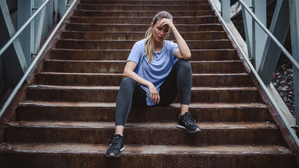 A woman sitting down on steps in workout clothes due to shortness of breath.