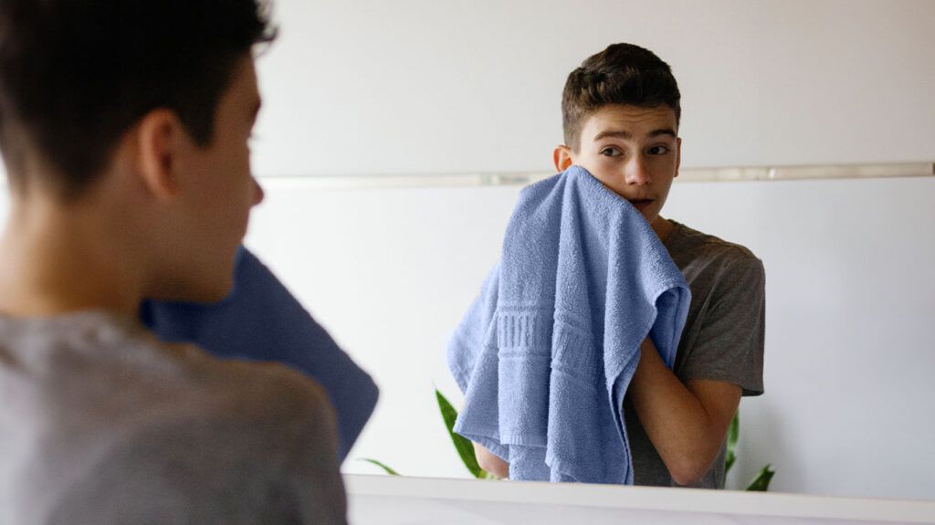 A teenage boy drying his face with a blue towel in front of a mirror during a skin care routine for acne.