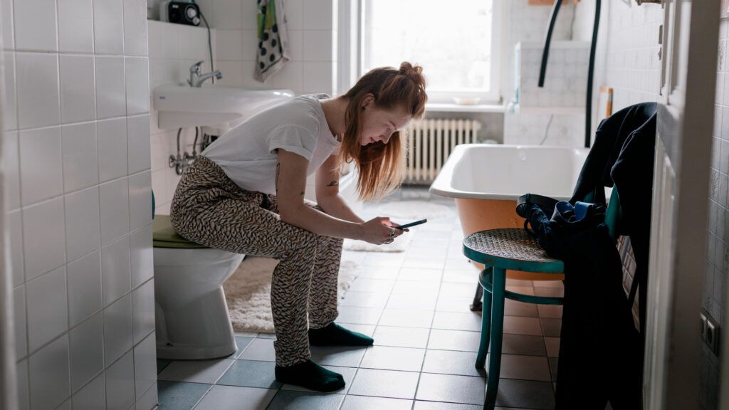 A woman sitting on a closed toilet while looking at her phone.