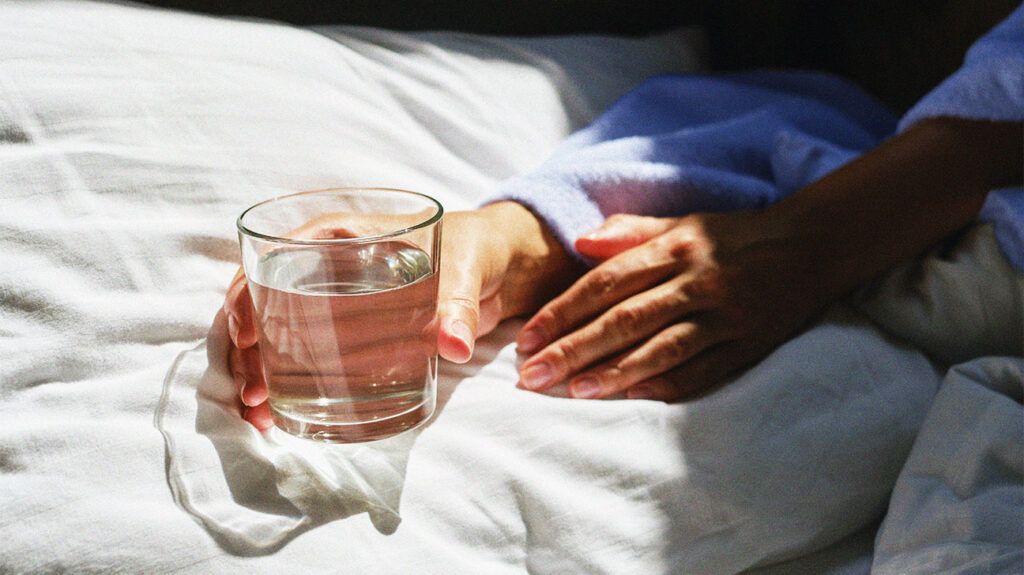 Close up of a pair of hands holding a glass of water, surrounded by linen bedsheets