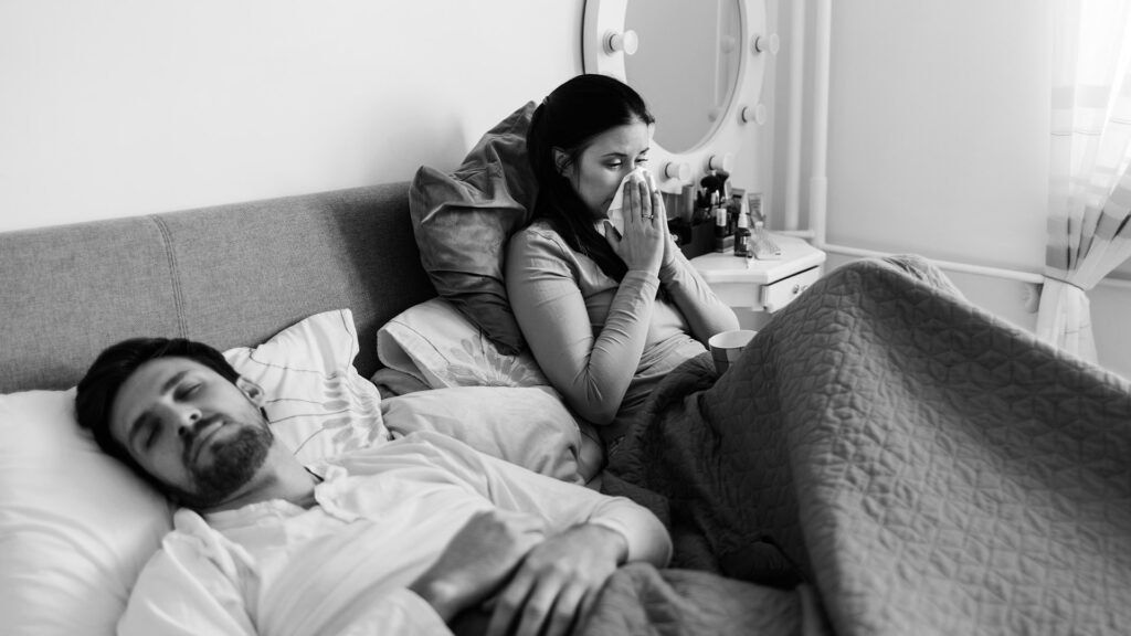 A woman sat in bed with mono next to her partner who is asleep.