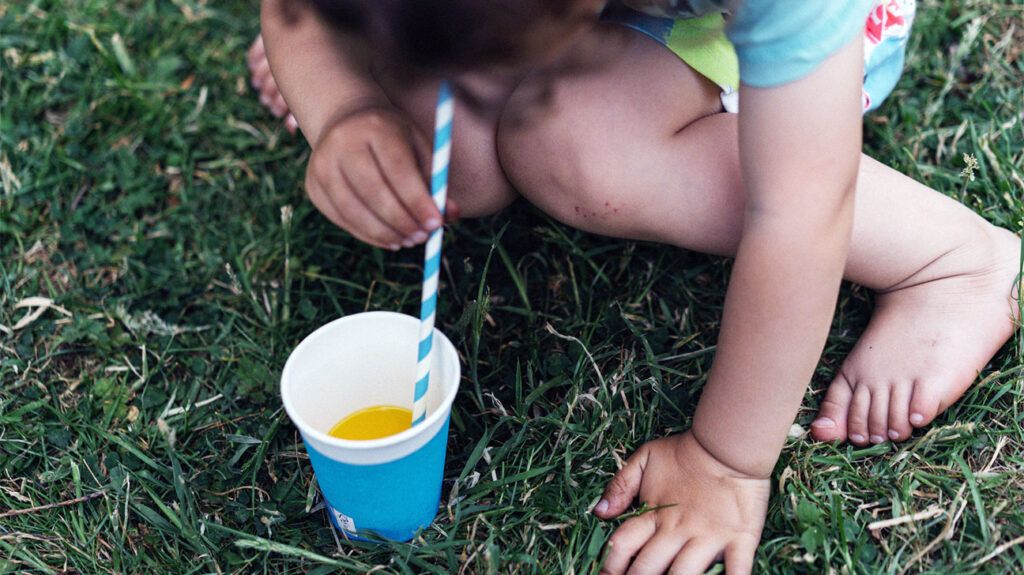 Primo piano di un bambino che beve succo d'arancia da una tazza usa e getta con una cannuccia sull'erba all'esterno