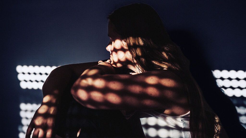 A woman sitting looking pensive in a dark room with dappled light. -1