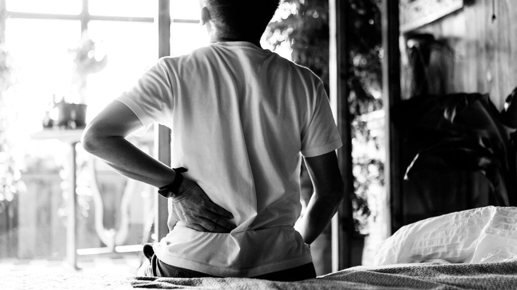 Black and white image of a male sitting on the edge of a bed holding his lower back