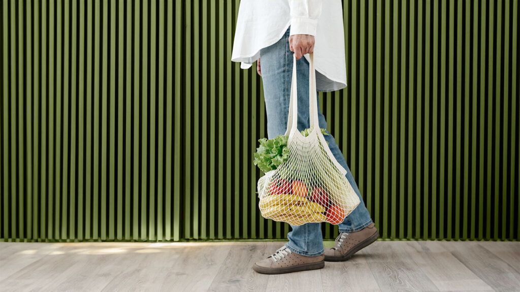 A close-up of a person carrying vegetables in a knit, reusable grocery bag.