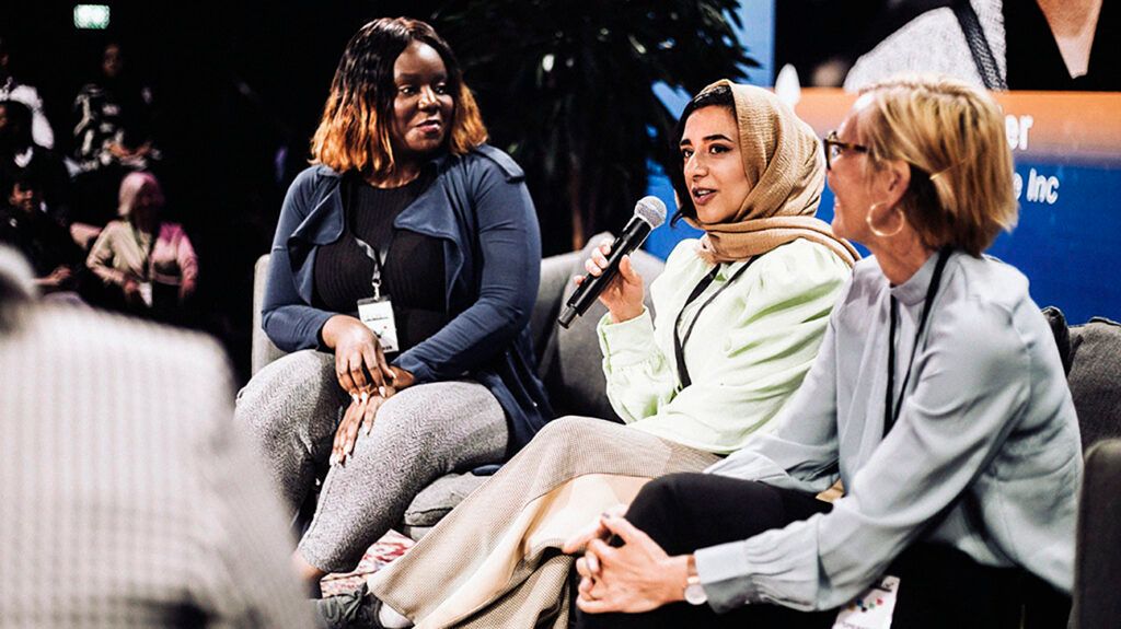 Three women sitting on a panel at a conference, one is talking into a microphone.