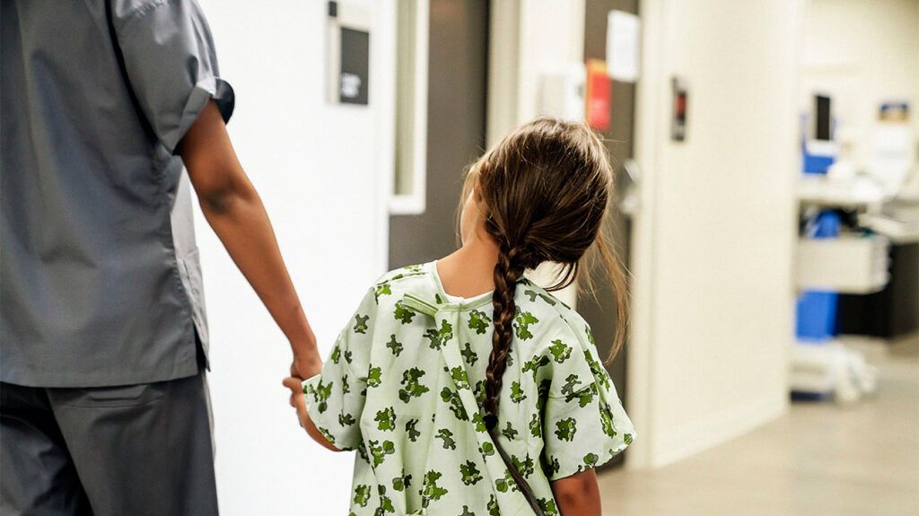 A child in a hospital gown is holding a medical professional's hand.