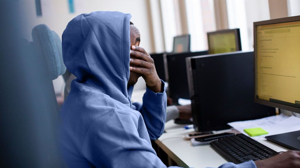 A man in a hoodie rubbing his dry eyes while using a computer.