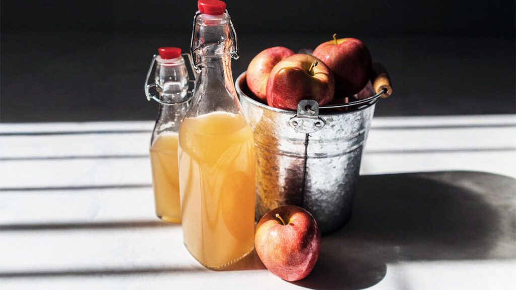 two bottles of apple cider vinegar next to a bucket of red apples