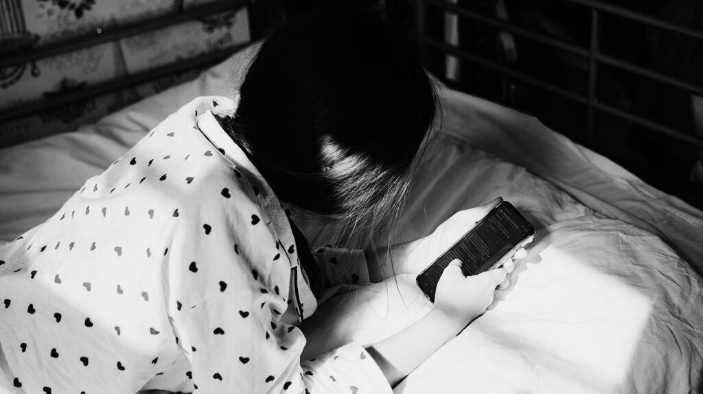 Black and white image of a teenager lying on a bed looking at their phone