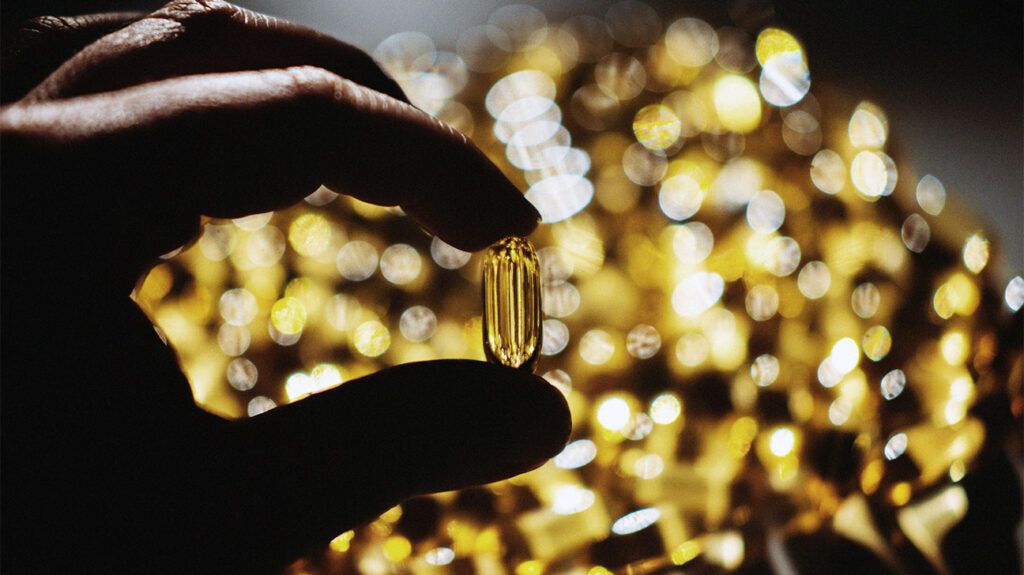 A hand holding a soft yellow capsule of vitamin D on a sunlit background of more capsules