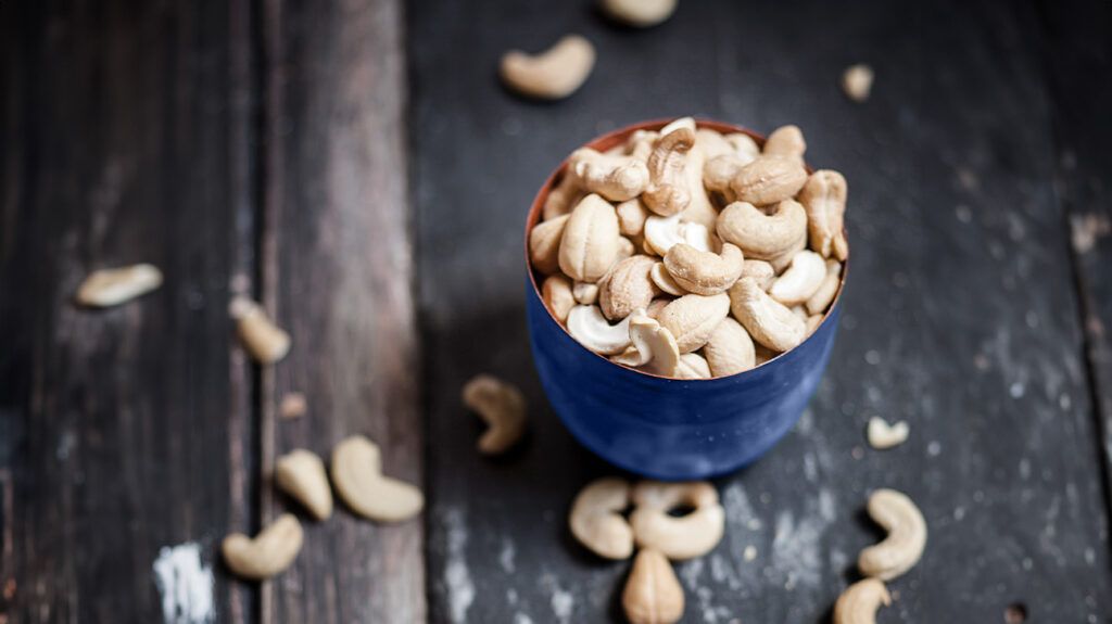 A cup full of cashews, with more cashews scattered around it, on a table.
