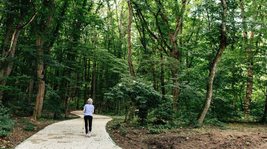 An older woman walks on a wide path through a forested area
