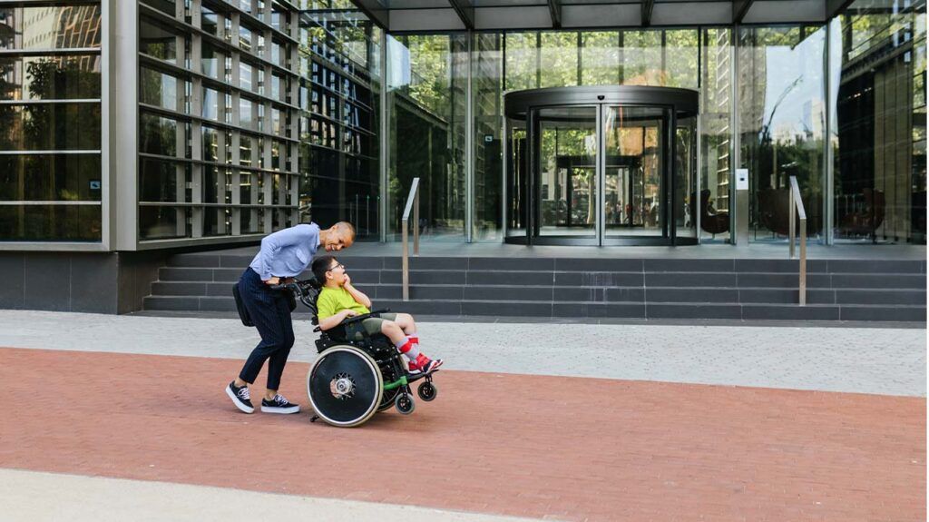 A child with muscular dystrophy in a wheelchair with their carer pushing them from behind.-1