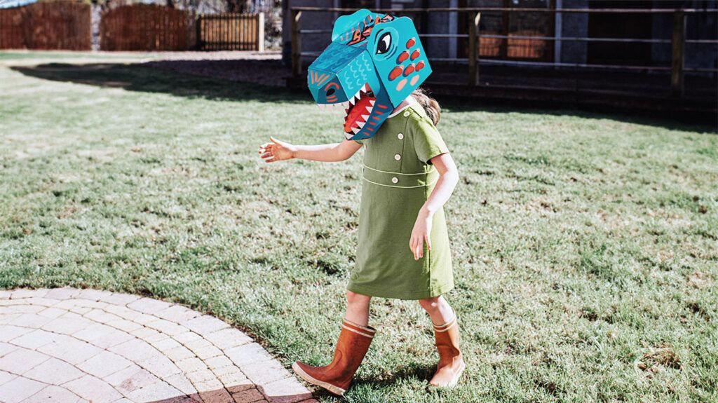 A girl in a garden wears a brightly colored cardboard dinosaur mask-1