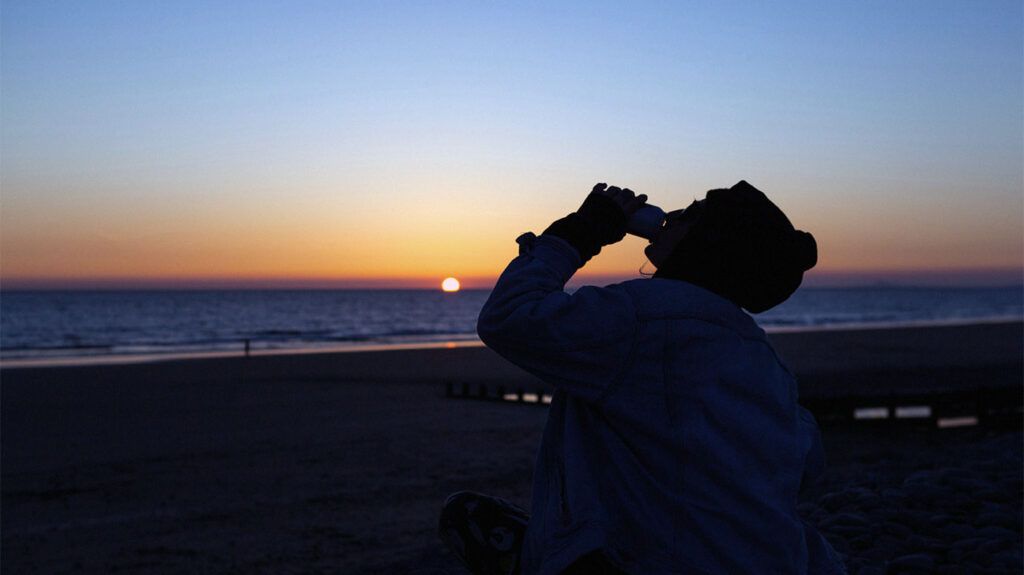 A person drinking a can of soda at sunset in this backlit image.