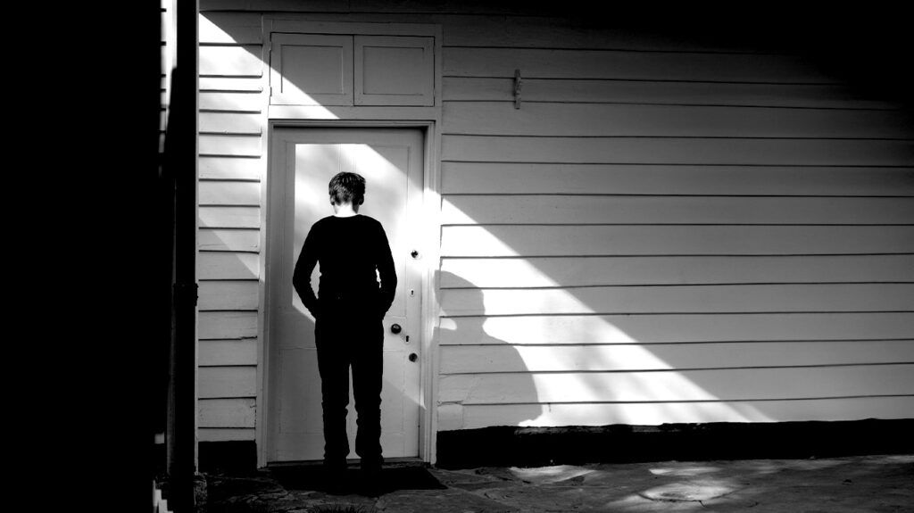 Black and white image of a young person standing in front of a closed door looking uncertain