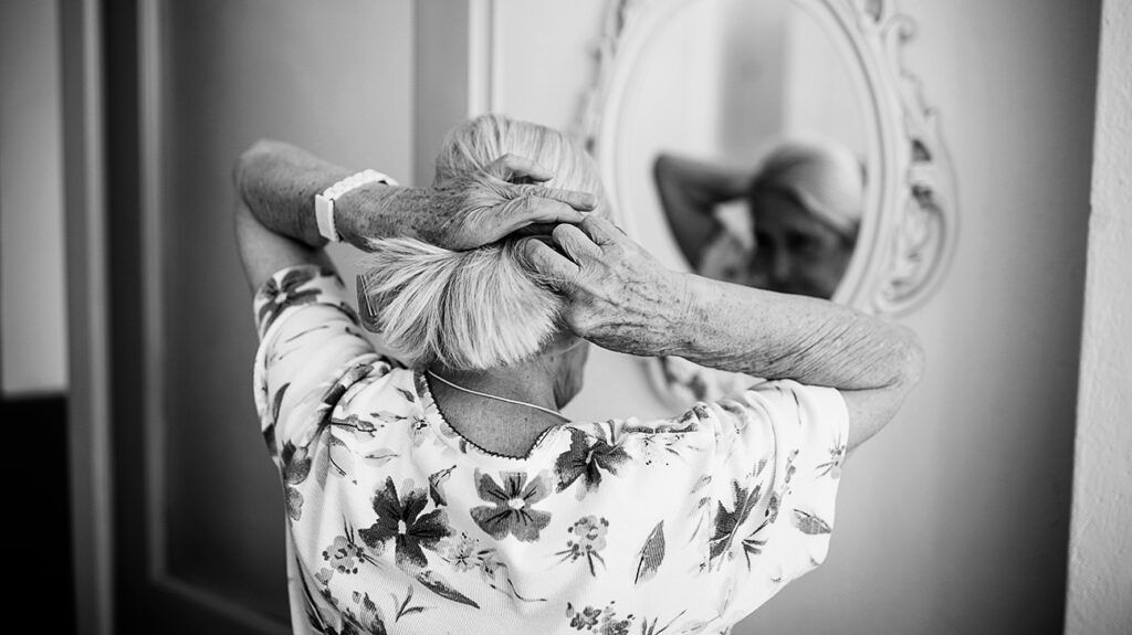 An older woman putting her hair in a ponytail in front of a mirror.