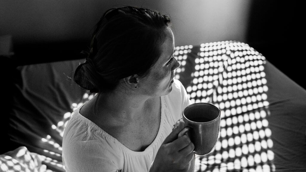 A woman drinking coffee while sat on a bed.