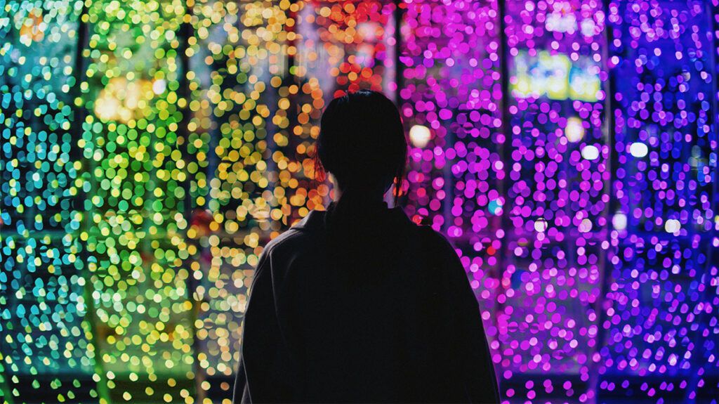The silhouette of a young woman standing against illuminated and colourful bokeh lights that look like beads.