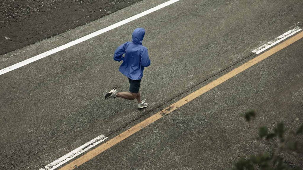 A man wearing a windbreaker runs in the middle of a road.