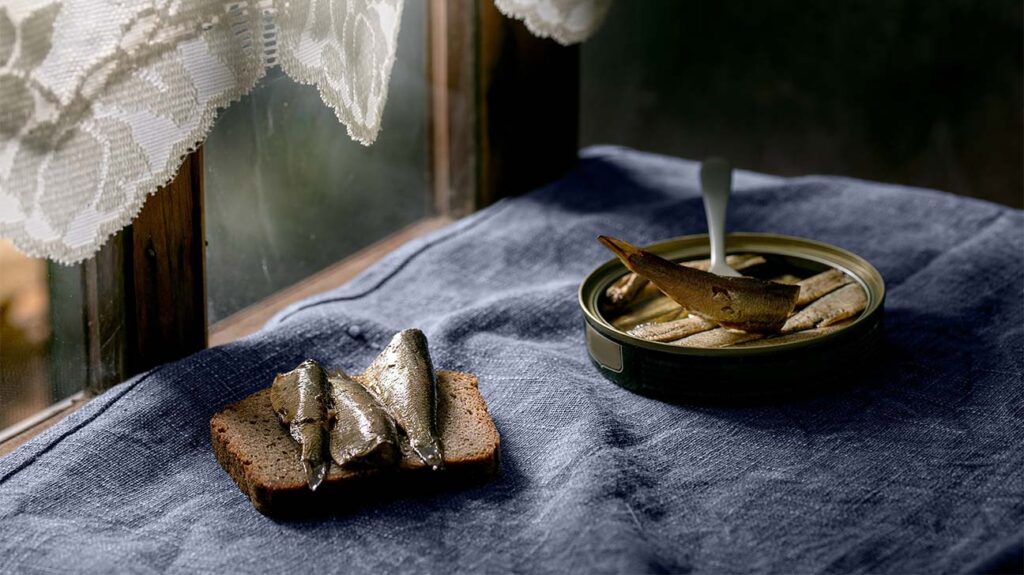 A can of sardines and some fish on a slice of bread by a window