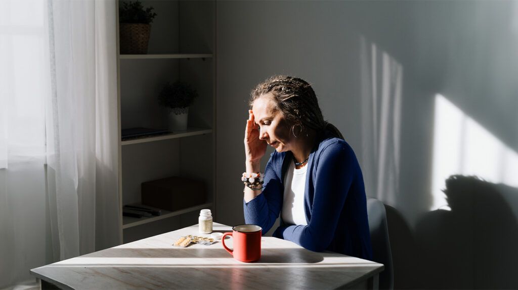 A woman with a migraine headache holds her head while sitting at a table
