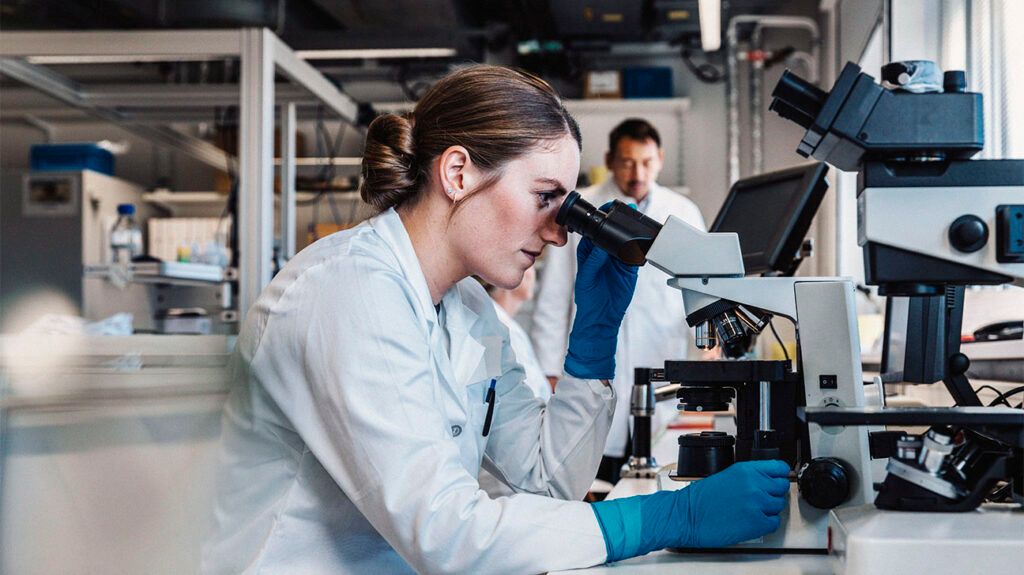 Young woman analyzing a slide under a microscope with a man in the background.